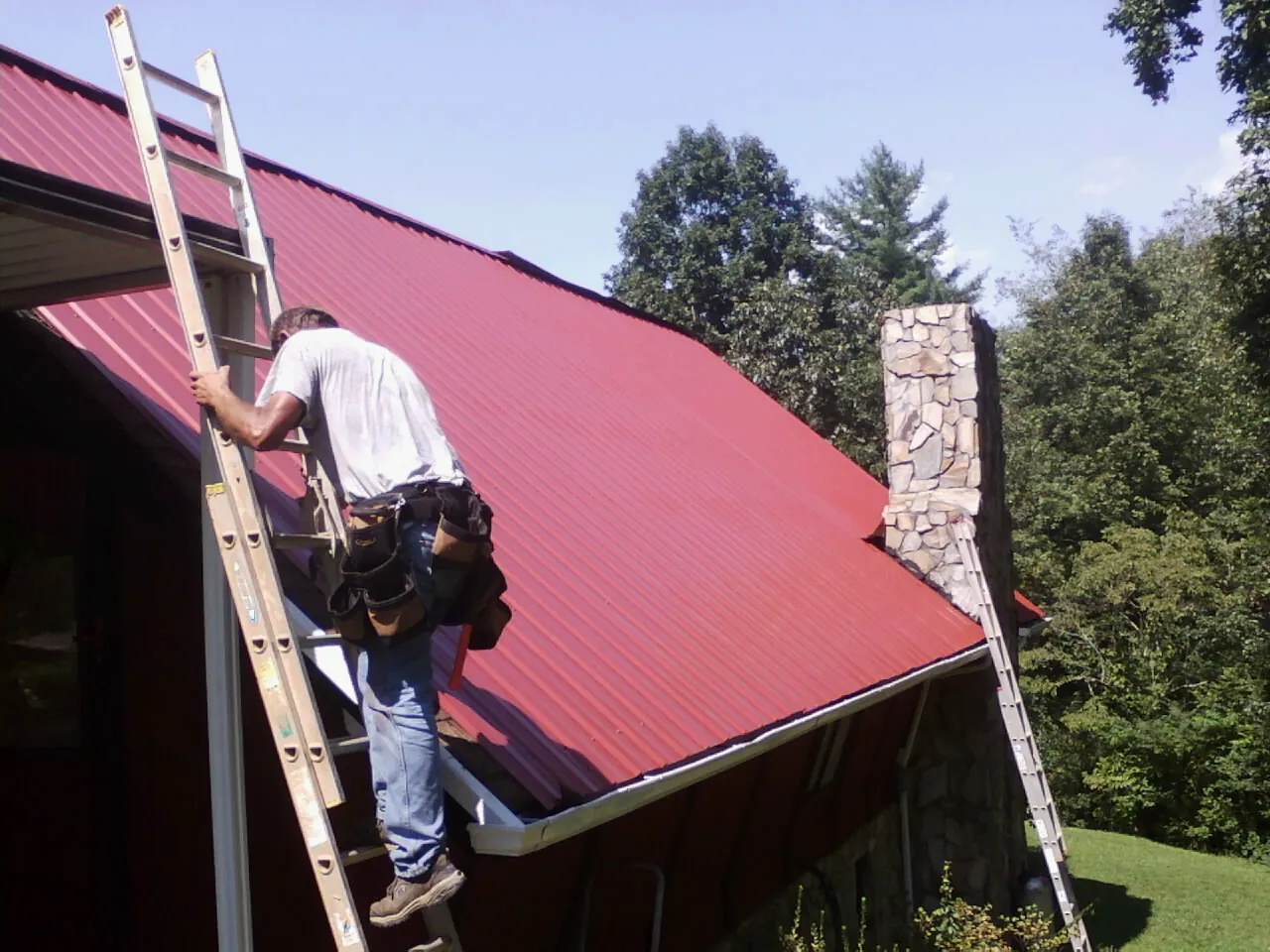 Roofer on a ladder beside a red metal roof and stone chimney — Soffit & Fascia Repair by Roofing Brothers in Georgiana